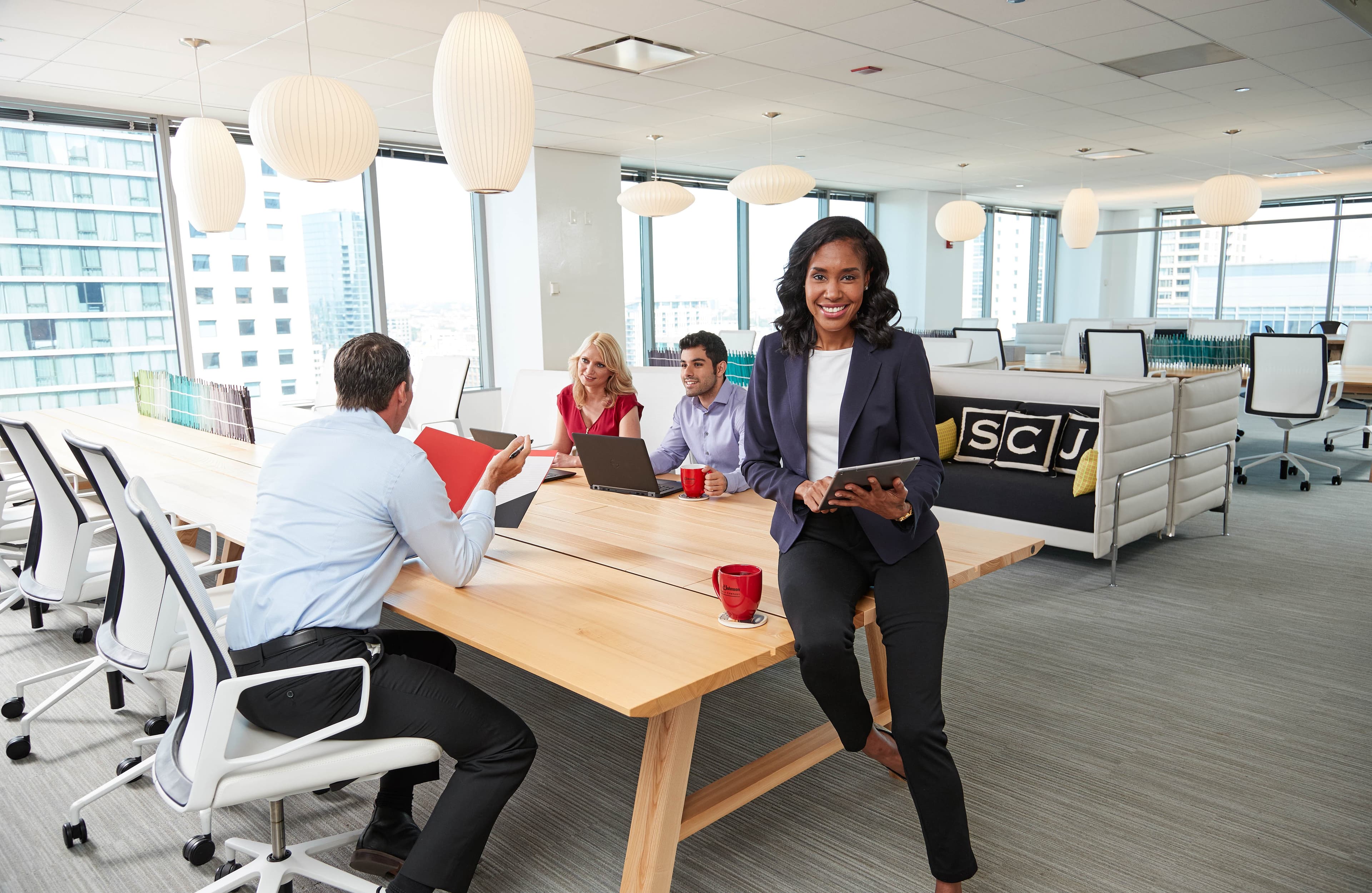 A person holds a tablet, smiling, and leaning on a table in a modern office. Three others sit at the table with laptops and red mugs. Cushions display "SCJ."