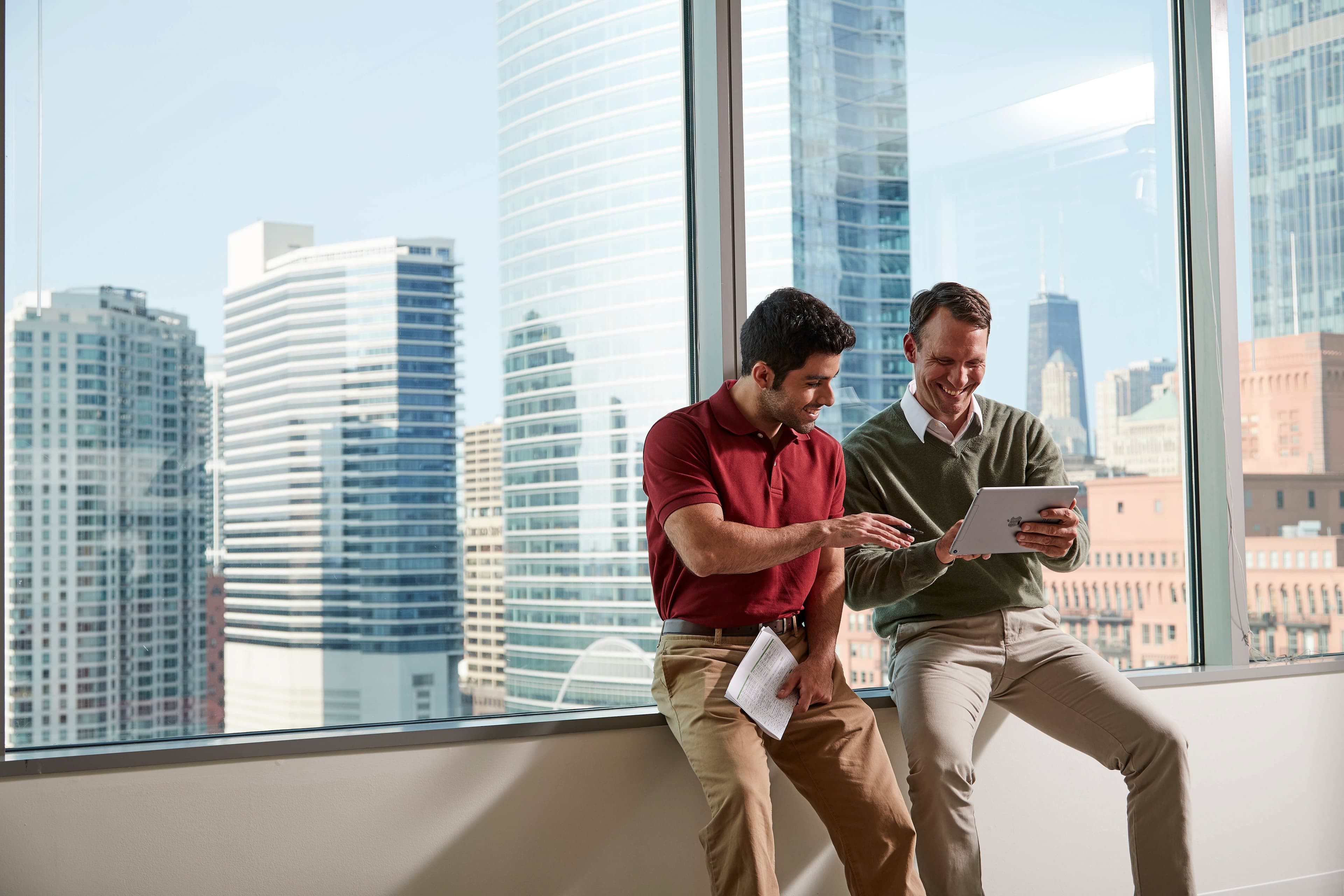 Two men sit on a windowsill, one holding a tablet and smiling, the other pointing at the screen. They are in a high-rise office with a city skyline view.