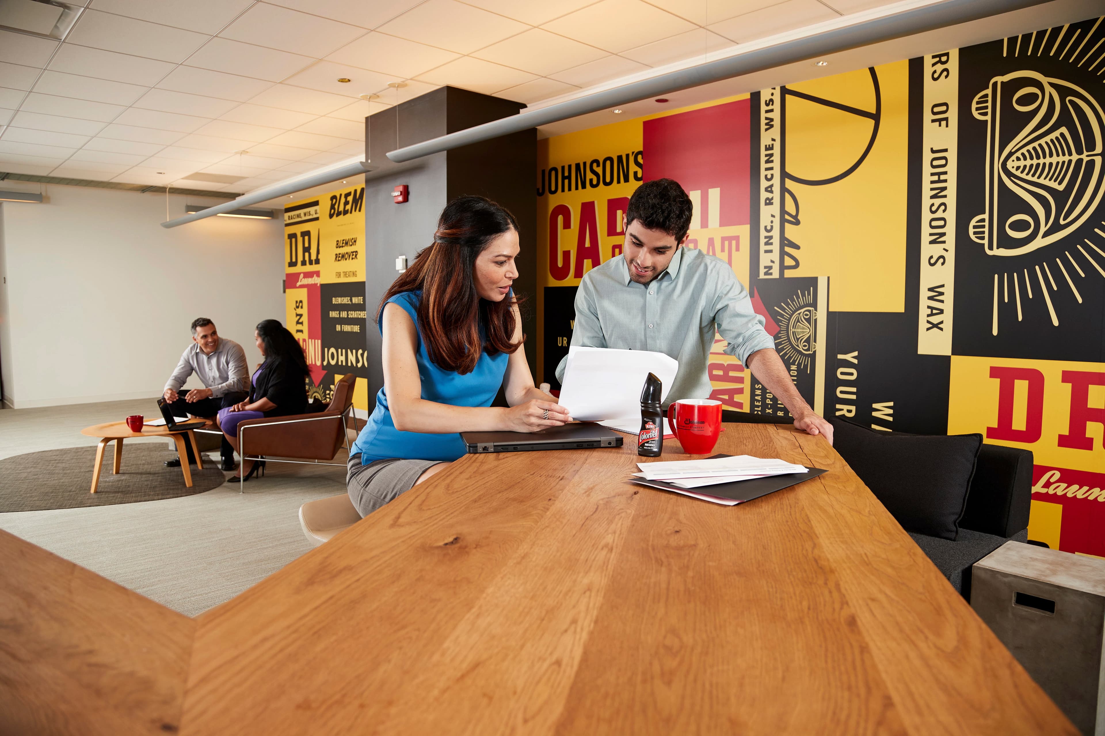 Two people review documents at a wooden table in a modern office. Wall displays large, colorful Johnson's Wax advertisements. In the background, two others converse on a sofa.