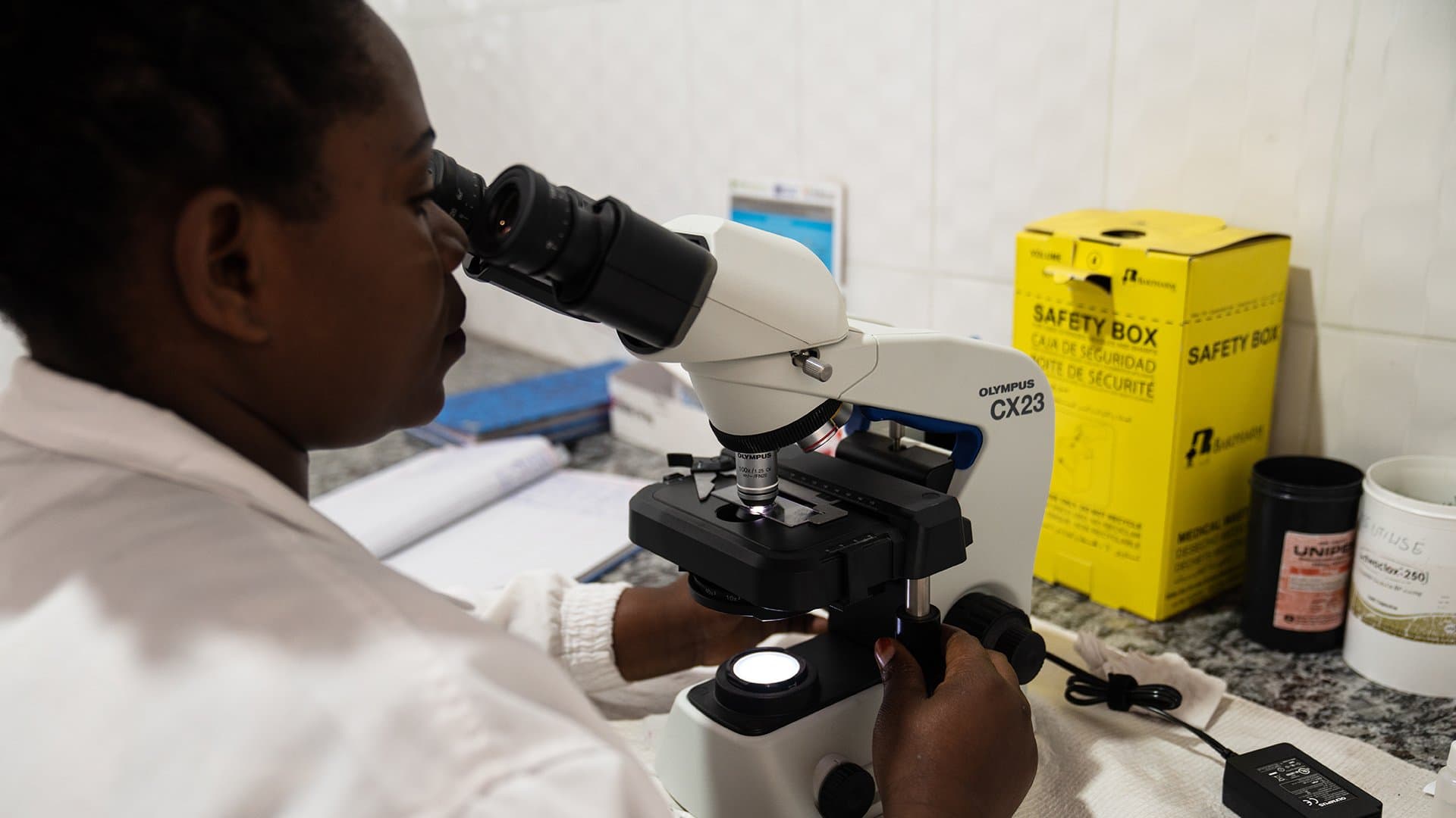 A person operates an Olympus CX23 microscope. In a laboratory setting, a yellow "SAFETY BOX" is visible nearby along with various containers and scattered papers.