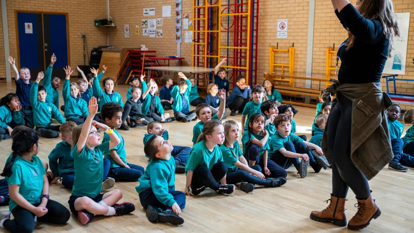 Children in teal uniforms are seated on a gym floor, eagerly raising their hands. A teacher, partially visible, stands in front, addressing them. The gym has stacked chairs and wall bars.
