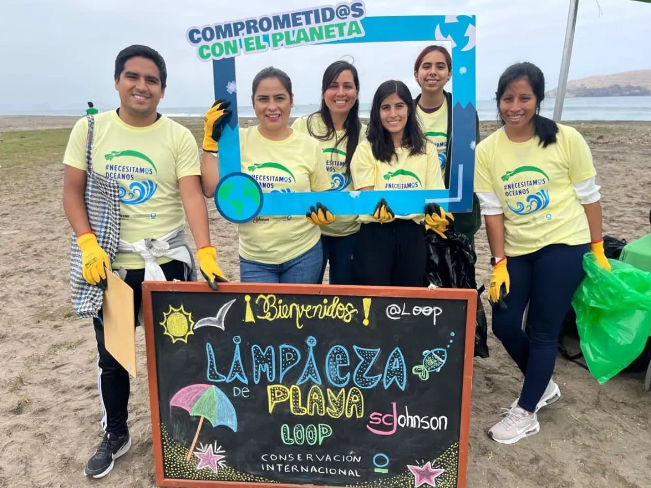 People wearing yellow shirts and gloves stand on a beach, holding a blue frame. They're next to a decorative chalkboard sign reading "¡Bienvenidos! Limpieza de Playa LOOP Conservación Internacional SC Johnson."
