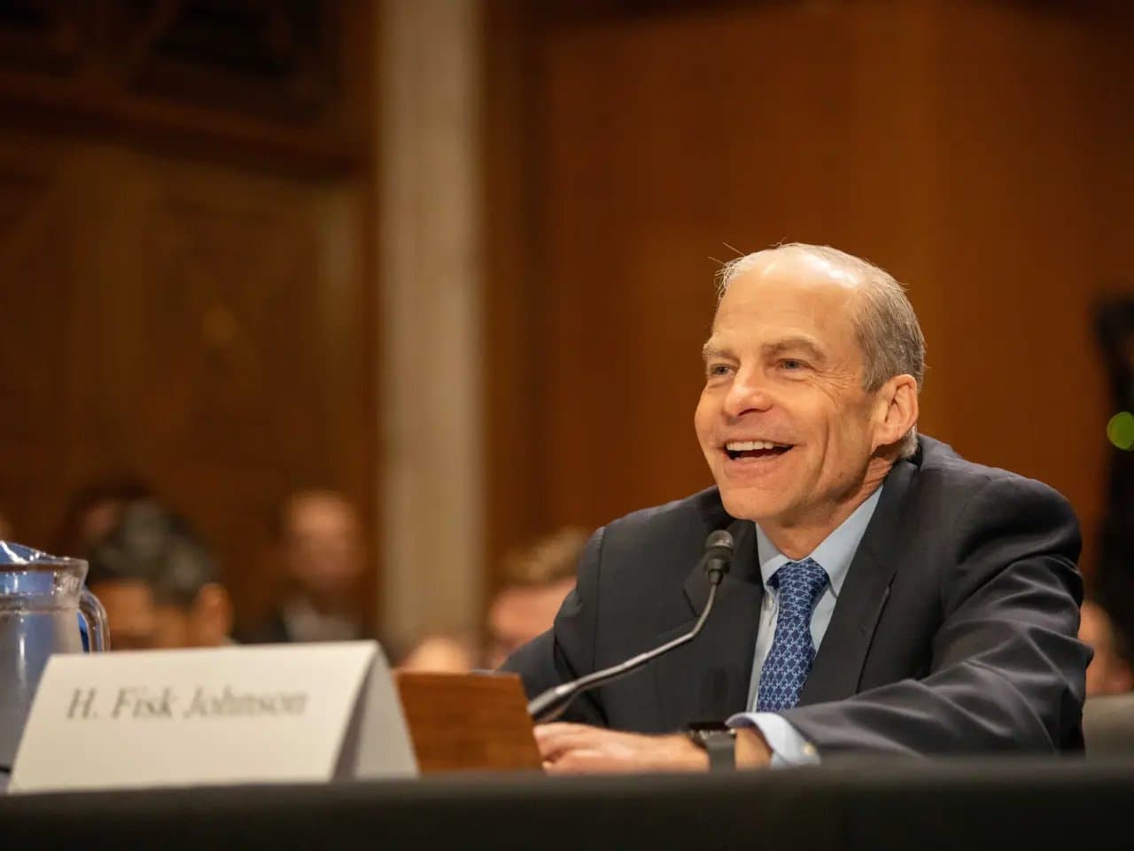 A man sits and speaks into a microphone, smiling, in a formal setting. A nameplate reads "H. Fisk Johnson." He is wearing a dark suit with a blue tie, surrounded by others.