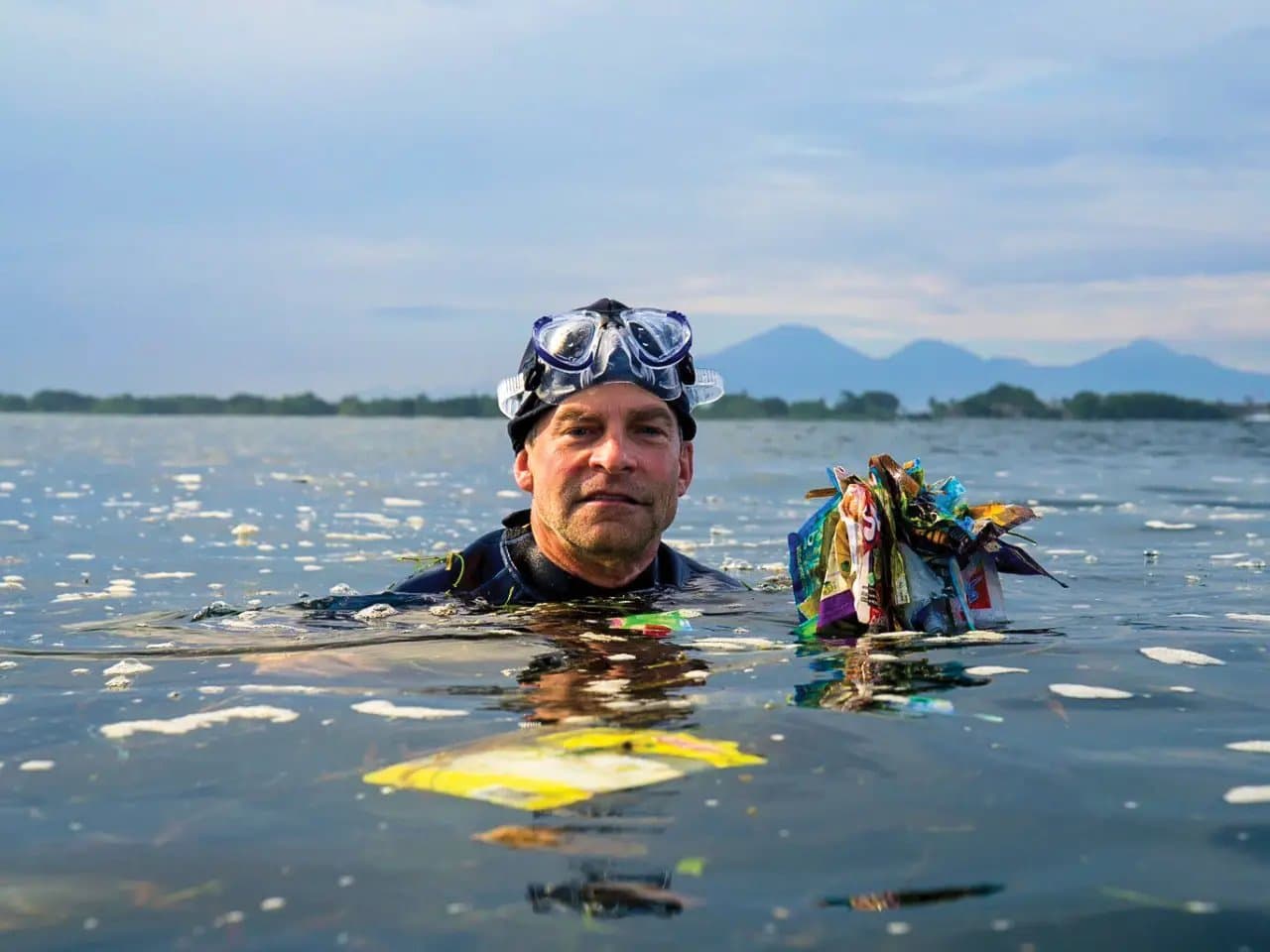 A diver, wearing goggles, is partially submerged in water, holding a bundle of colorful trash. The background shows a distant shoreline and mountains under a cloudy sky.