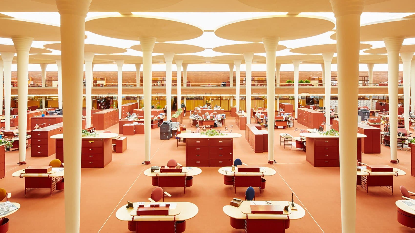Desks and cabinets fill a large, open office space featuring columns with circular tops. Natural light filters through the ceiling openings, adding warmth to the organized, modern environment.