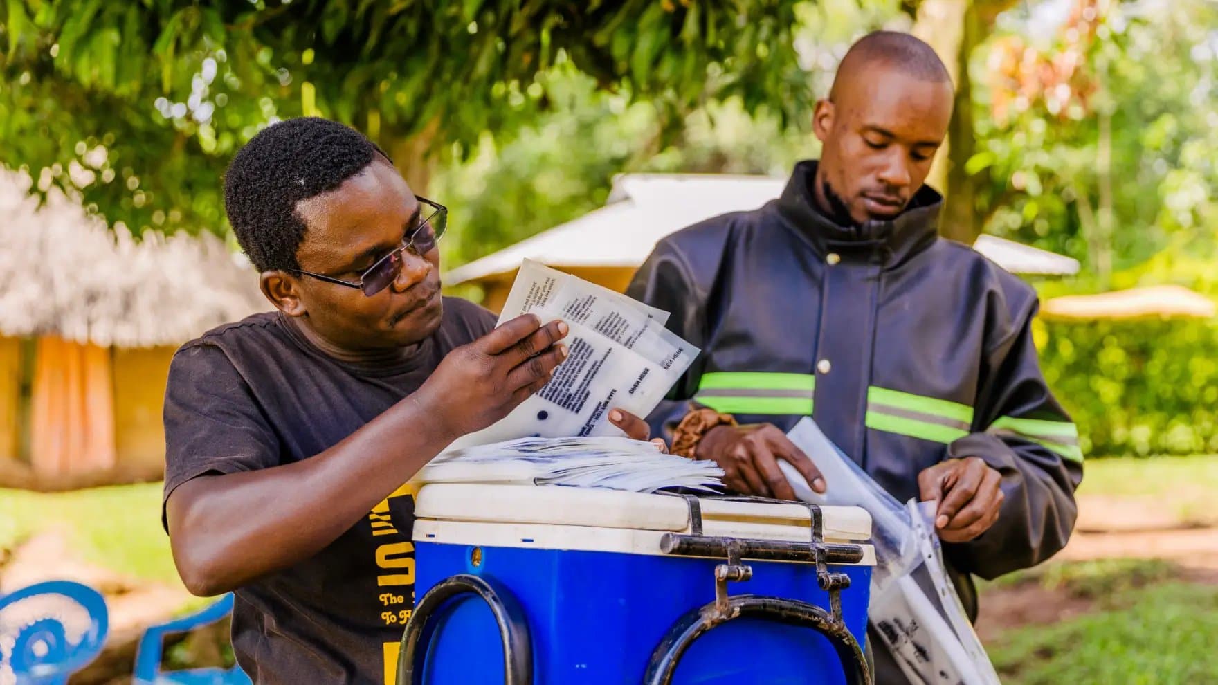 Two individuals handle packages near a large blue container, outdoors with green trees in the background. The packages appear to contain text, though specifics are not visible.