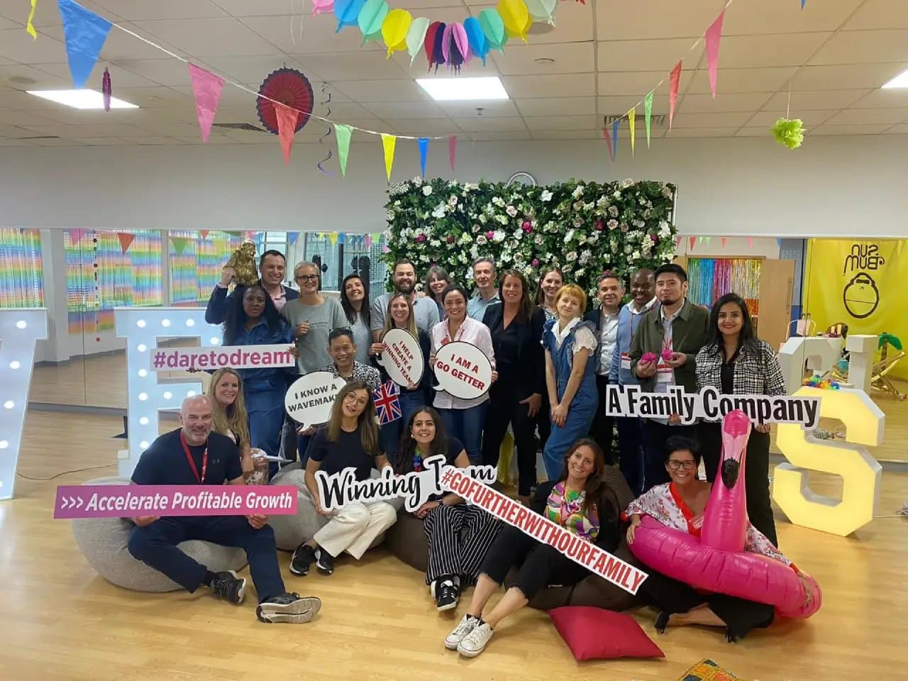 A group of people posing indoors, holding signs with phrases like "#daretodream," "A Family Company," "Winning Team," and "GO FURTHER WITH OUR FAMILY," in front of a floral backdrop and colorful decorations.
