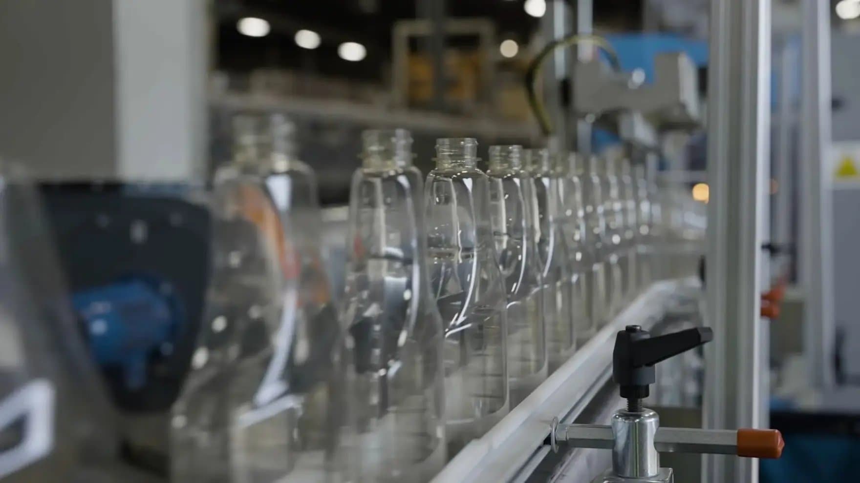 Clear plastic bottles are aligned on a conveyor belt, moving through an industrial setting with machinery and equipment in the background.