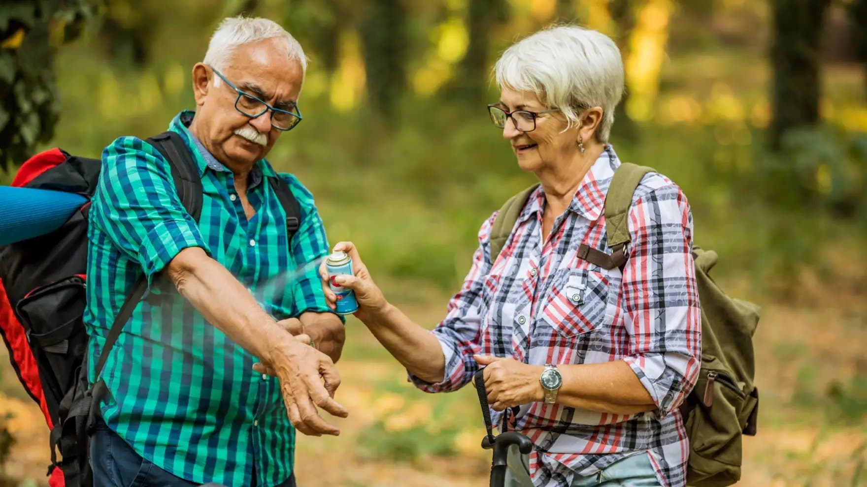 An elderly woman sprays insect repellent on an elderly man's arm in a forest setting. Both wear plaid shirts and backpacks, indicating a hiking activity.