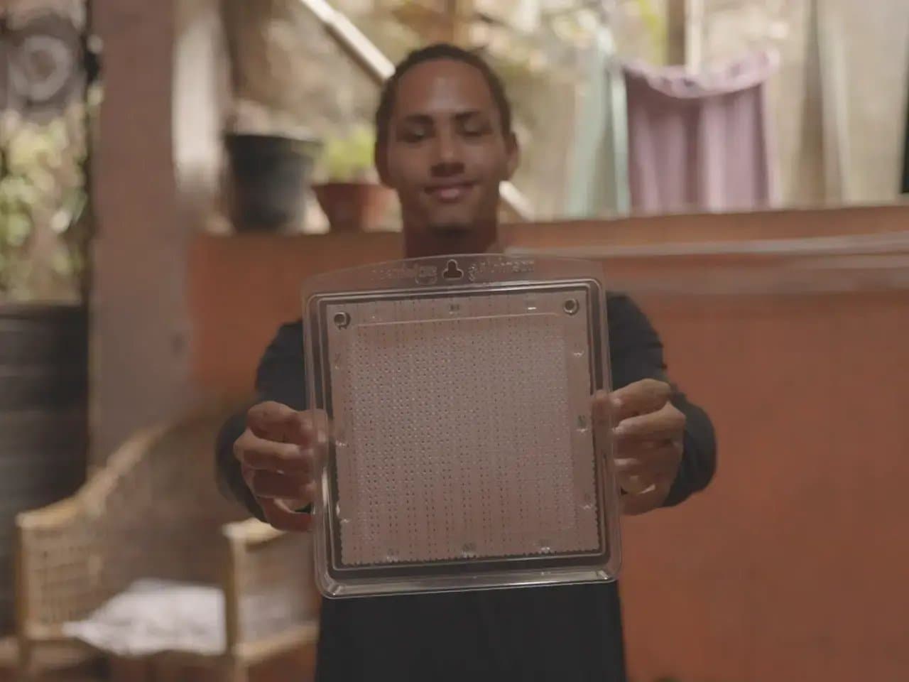 A person holds a clear square panel with a grid pattern, standing in an indoor setting with wicker furniture and hanging clothes in the background.