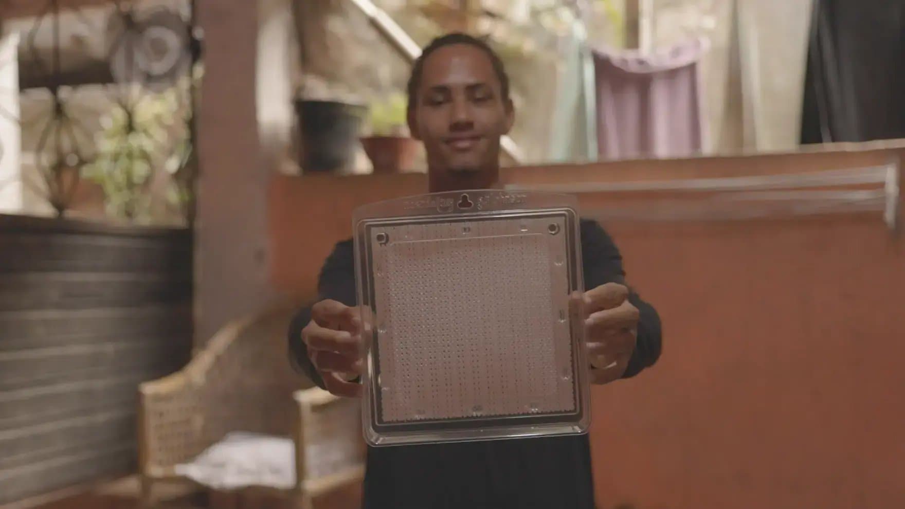 A person holds a transparent square object with perforations, smiling. The setting appears to be an indoor area with plants and furniture visible in the background.