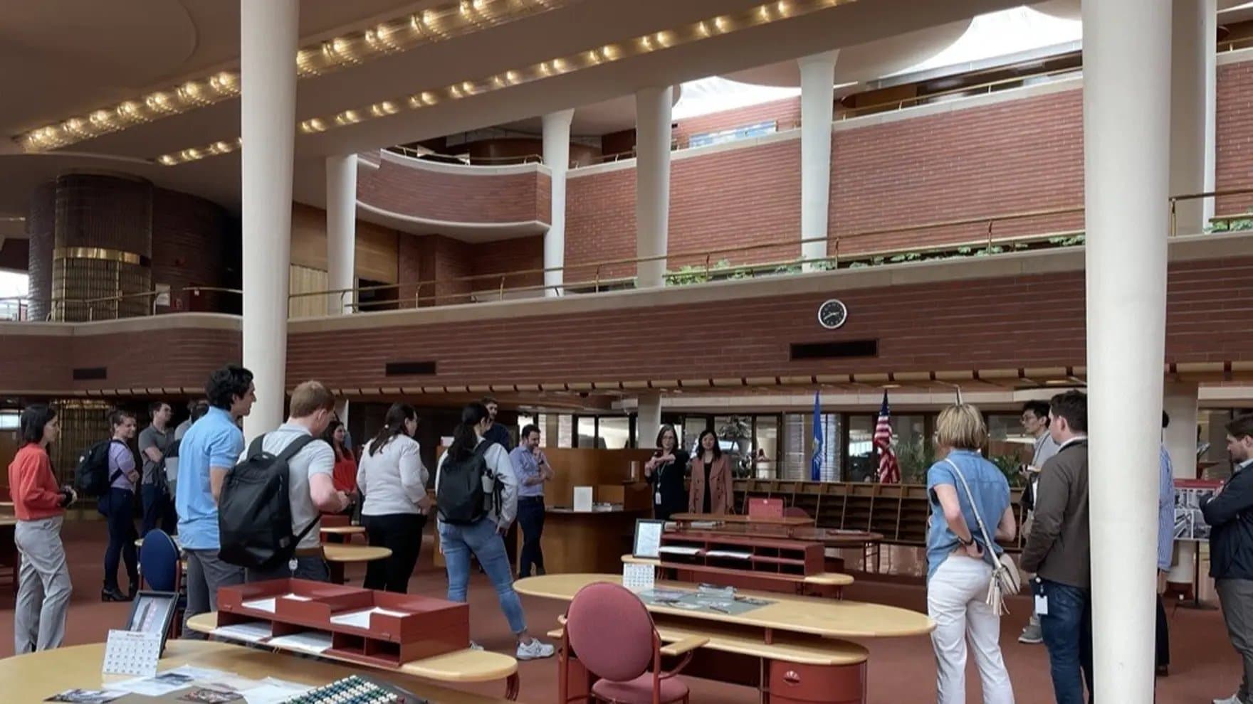 People gather in a large, modern office space with round desks and red chairs. The area has high ceilings, large white pillars, brick walls, and a clock on the wall.