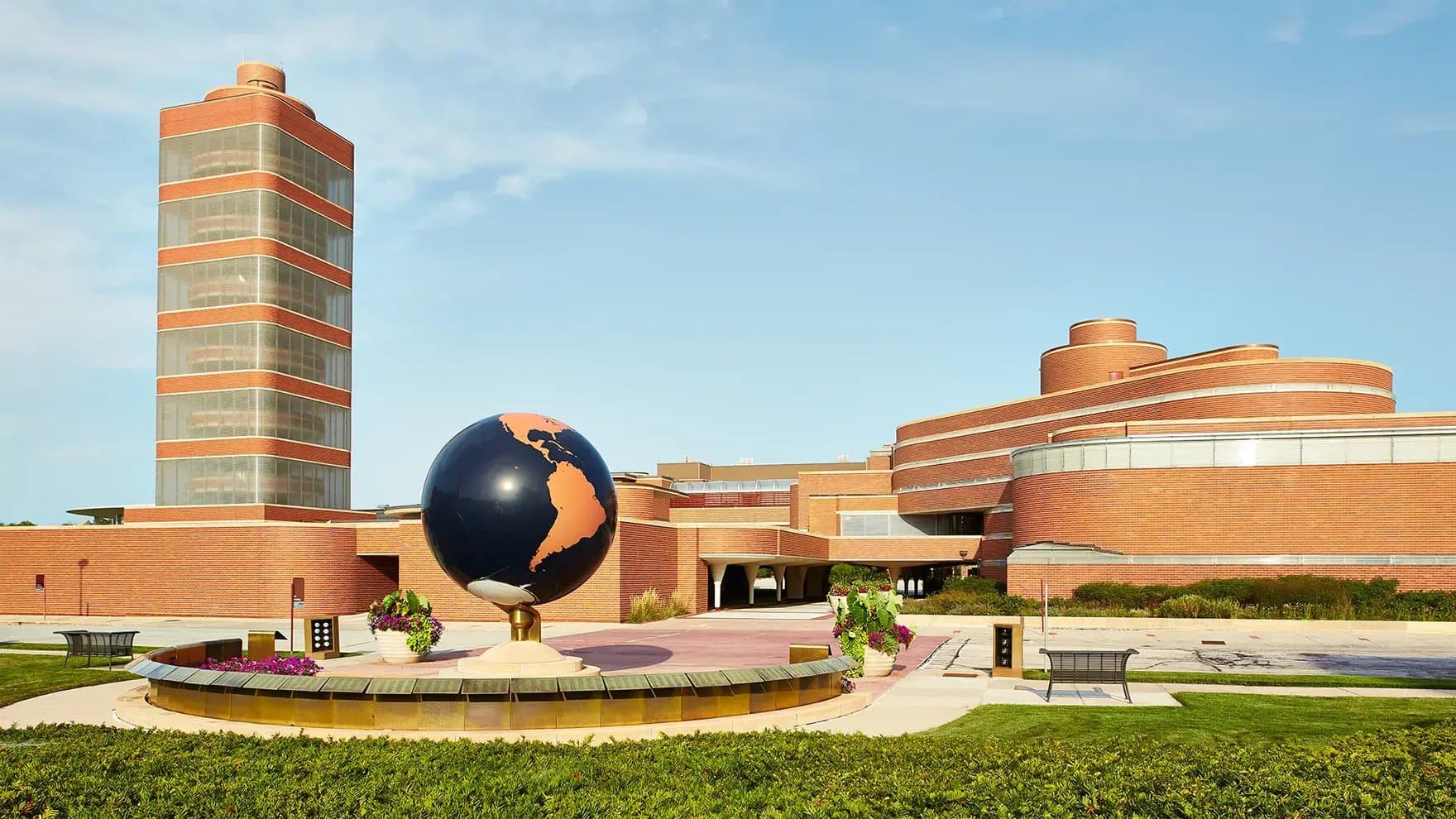 A large globe sculpture stands on a pedestal, surrounded by planters with flowers. It is set in front of a modern, red-brick building with circular and rectangular sections.