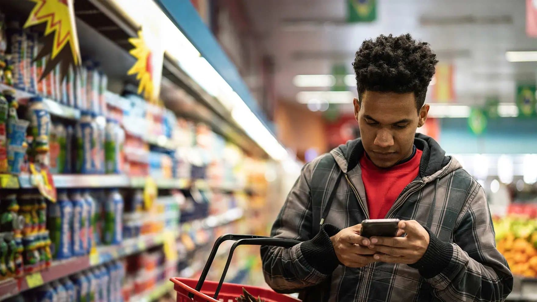 A person checks their phone while holding a shopping basket in a grocery store aisle filled with various products. Bright lights illuminate the shelves, creating a colorful ambiance.