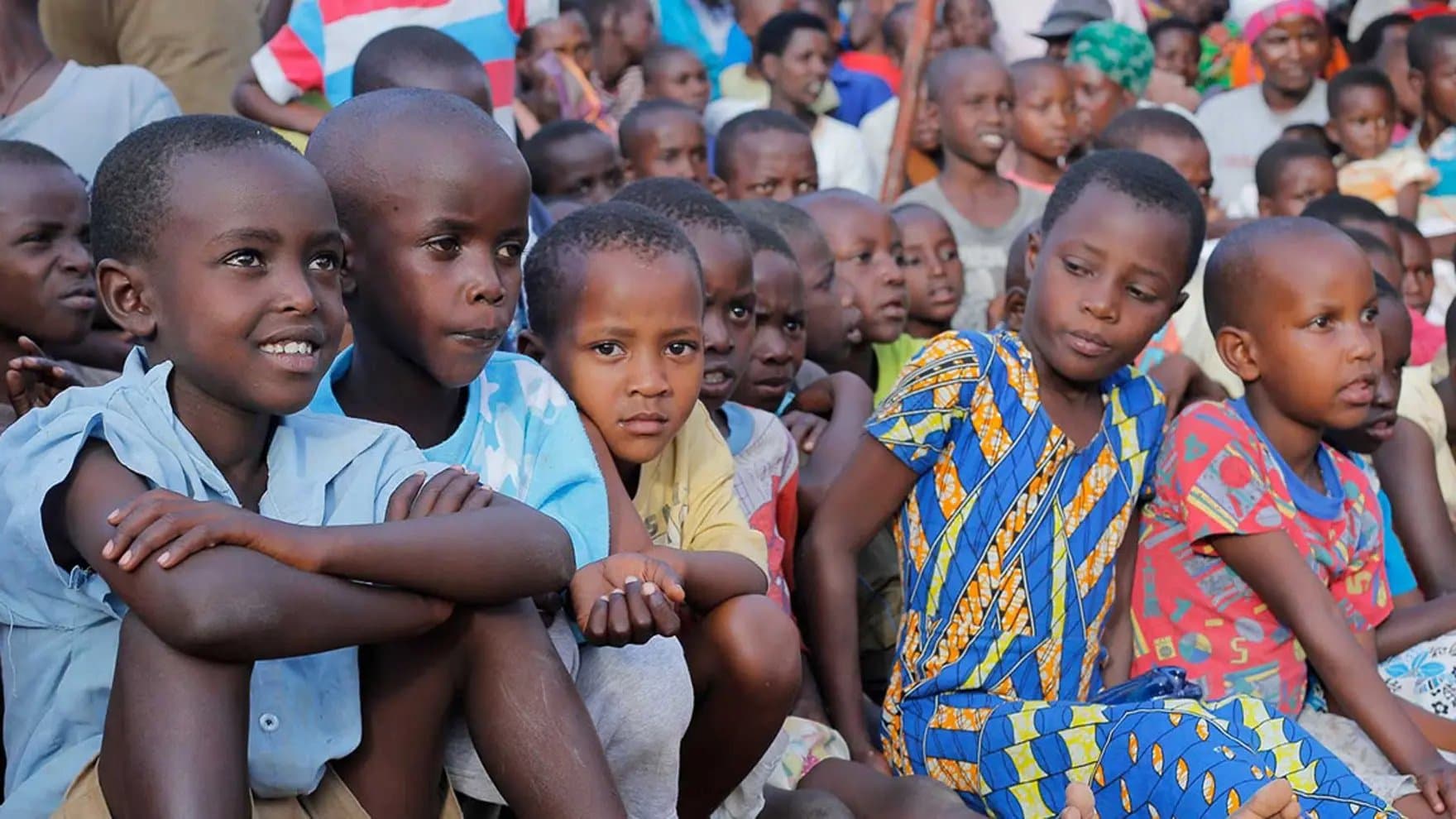 Children are sitting closely together, some smiling, while others look curiously or attentively at something. They wear colorful clothing, and the setting is crowded, suggesting a communal gathering or event.