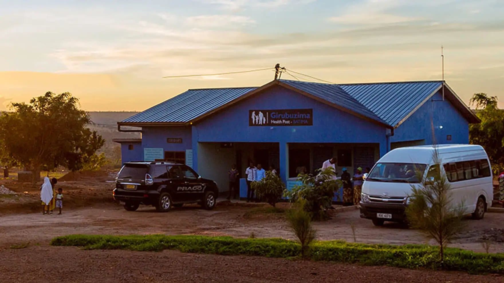 A blue building labeled "Girubuzima Health Post - SAKITA" sits with parked vehicles outside. People gather near the entrance under a sunset sky, surrounded by trees and dirt pathways.