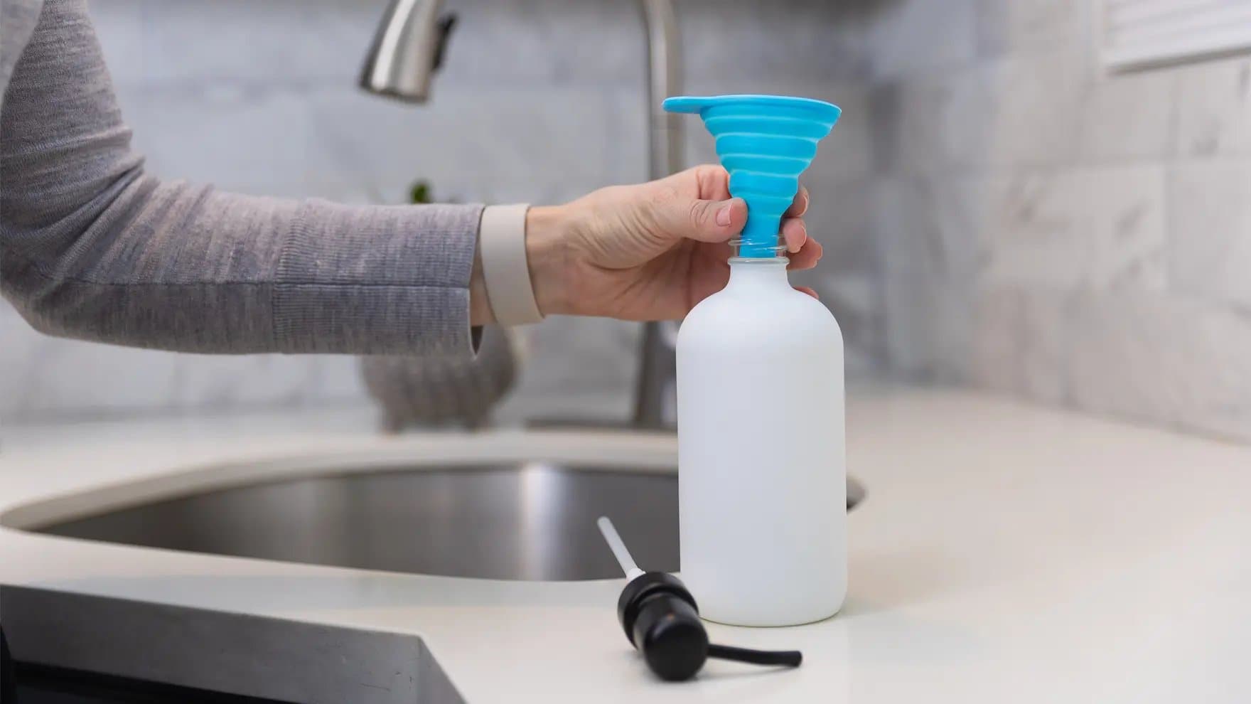 A hand holds a blue collapsible funnel over a white bottle on a kitchen counter near a sink, with a black pump lying nearby.