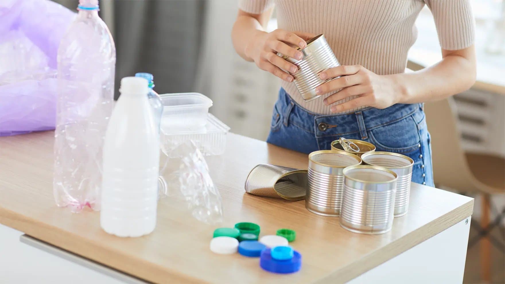 A person sorts metal cans on a wooden table, surrounded by plastic bottles, containers, and multicolored bottle caps, indoors.