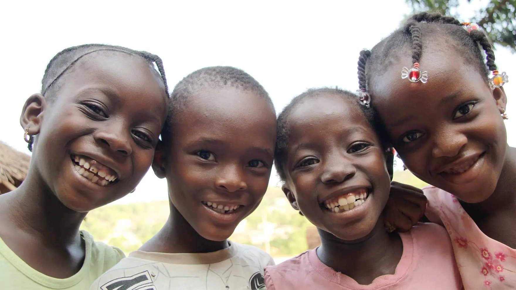 Four children smile joyfully at the camera, standing close together outdoors with trees and a clear sky in the background.