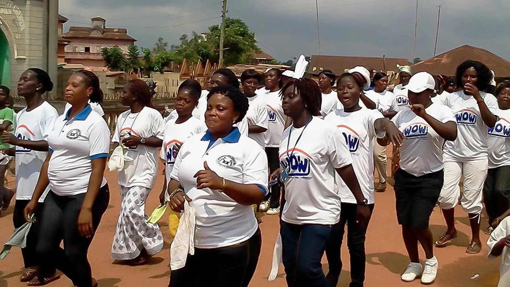 Participants march wearing white "WOW" t-shirts, holding cloths; they appear in an outdoor community setting, surrounded by buildings and greenery, during a daytime event.