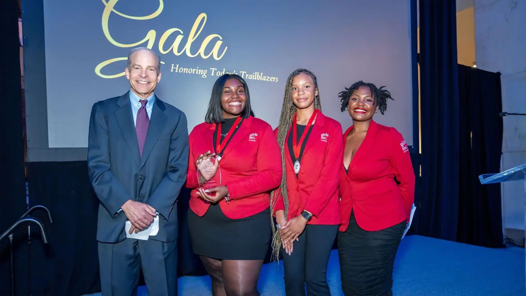 Four people stand on stage, three women in red blazers hold a trophy, one man in a suit smiles. Background text: "Gala Honoring Today's Trailblazers."