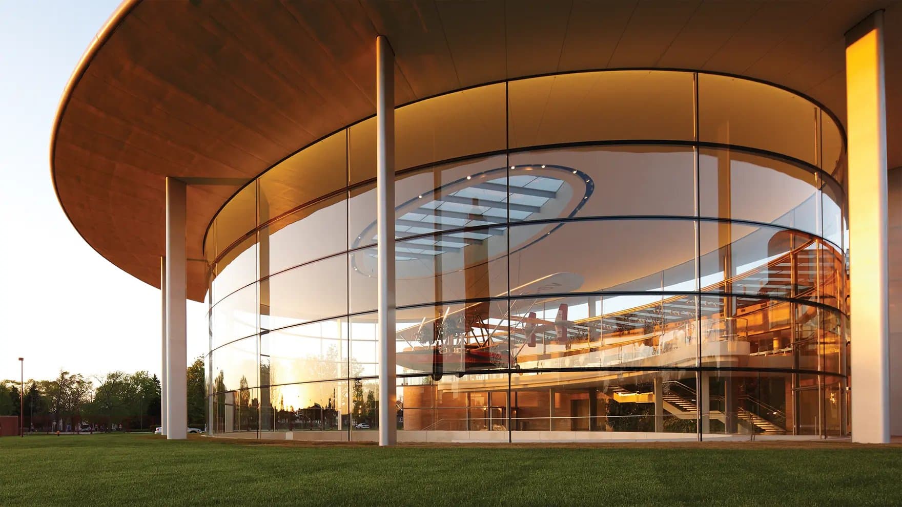 A modern, circular glass building reflects the sunset, showcasing sleek architectural lines. Inside, a vintage airplane is displayed prominently. The surrounding area features a well-manicured lawn.