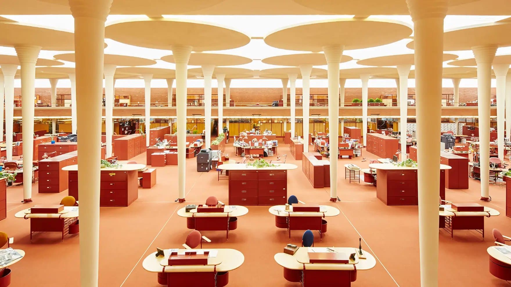Large open-plan office features red desks and chairs, surrounded by tall, white columns. The room is spacious with a warm color palette, creating a vibrant, organized workspace.