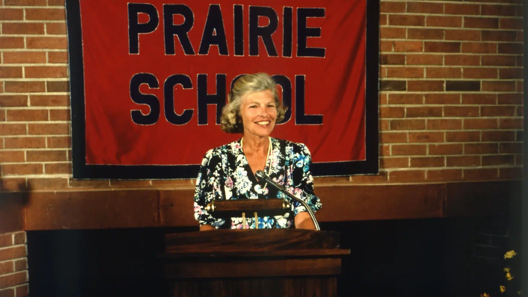A woman stands smiling at a podium with a microphone, against a red banner reading "PRAIRIE SCHOOL" on a brick wall background.