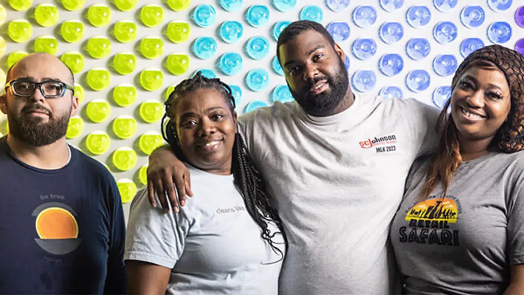 Four people stand closely together, smiling in front of a colorful wall with yellow, blue, and purple circles. Their shirts display "be true," "choose kindness," "SC Johnson MLK 2023," and "Retail Safari."