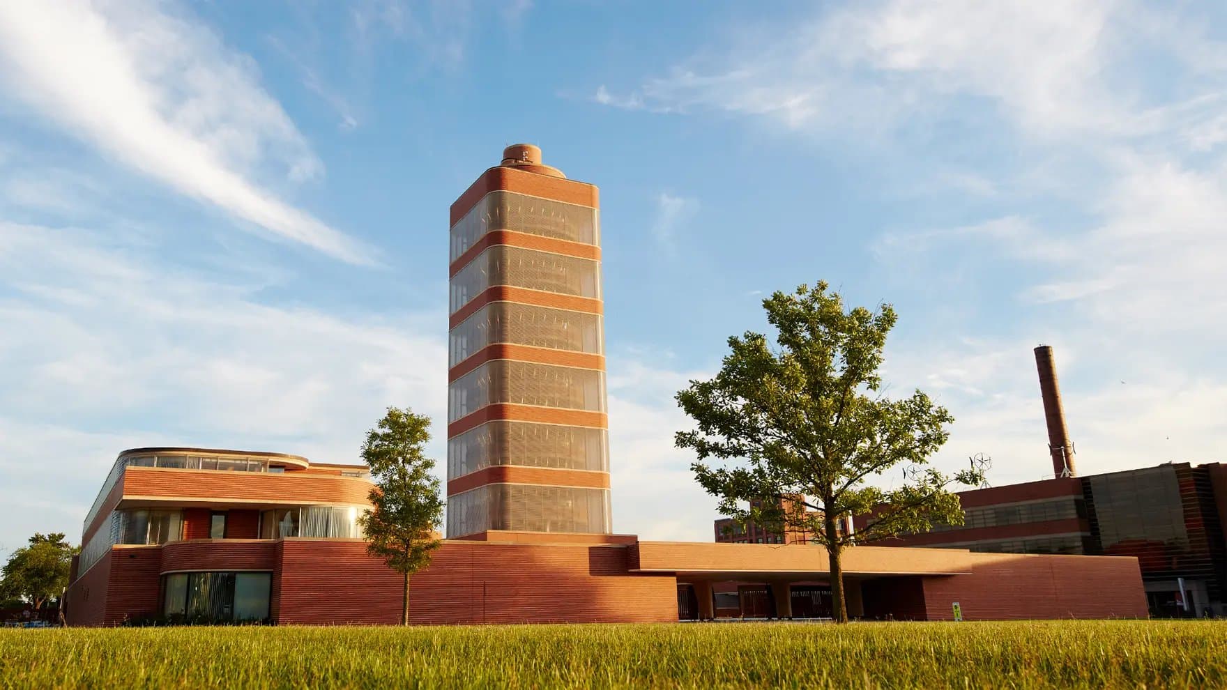 A tall, circular, multi-story brick building with wide bands stands in a green area, surrounded by grass and trees, under a blue sky. A factory-like structure is in the background.