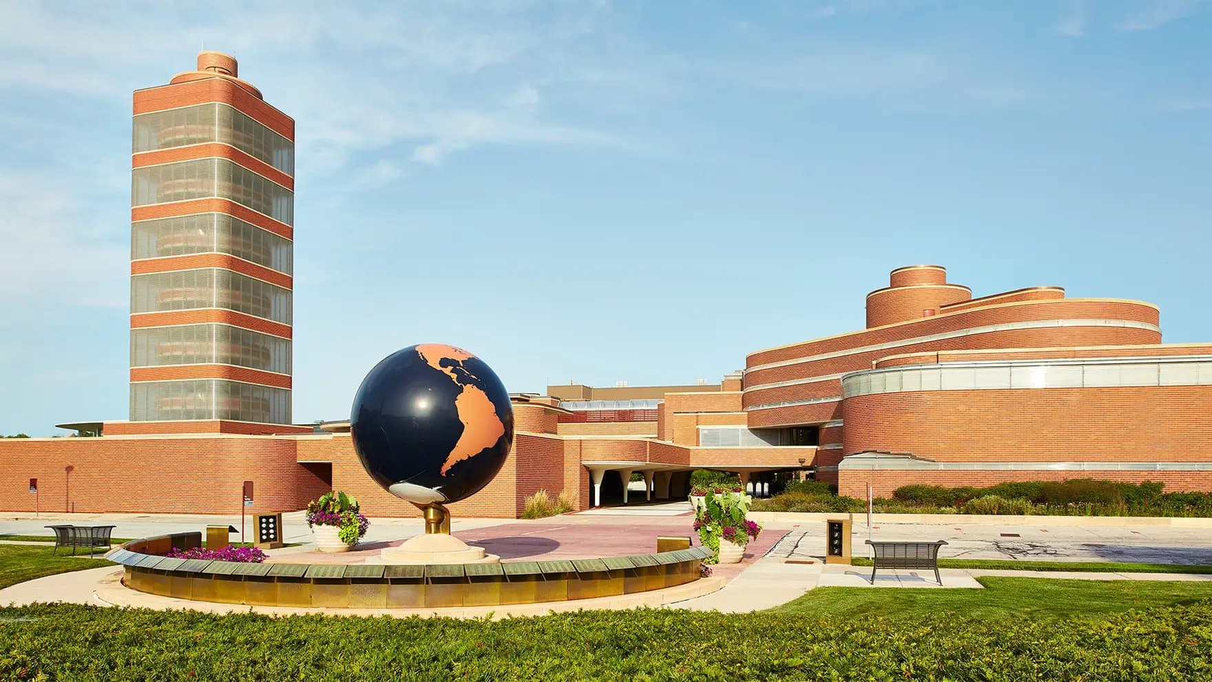 A large, blue globe statue stands prominently in front of a modern, red-brick building with multiple circular and rectangular structures, situated in a landscaped area with flowers and benches.