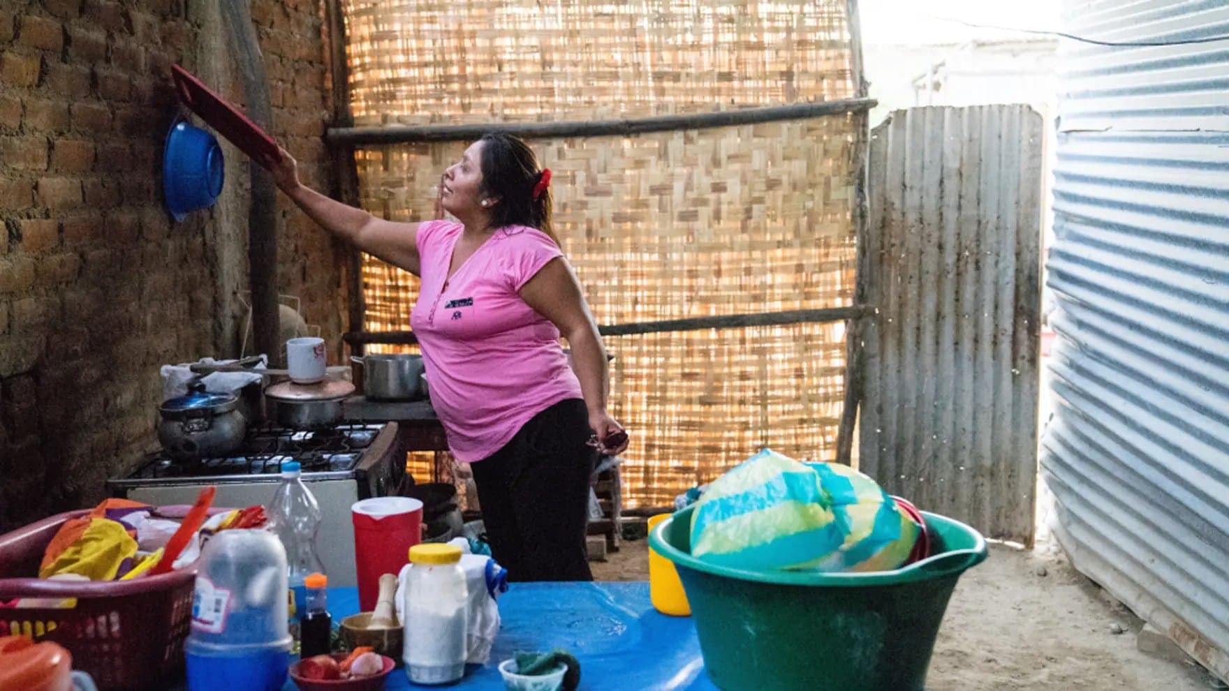 A woman in a pink shirt reaches up in a rustic kitchen. Pots are on a stove, and various containers sit on a table. The background features brick and woven walls.