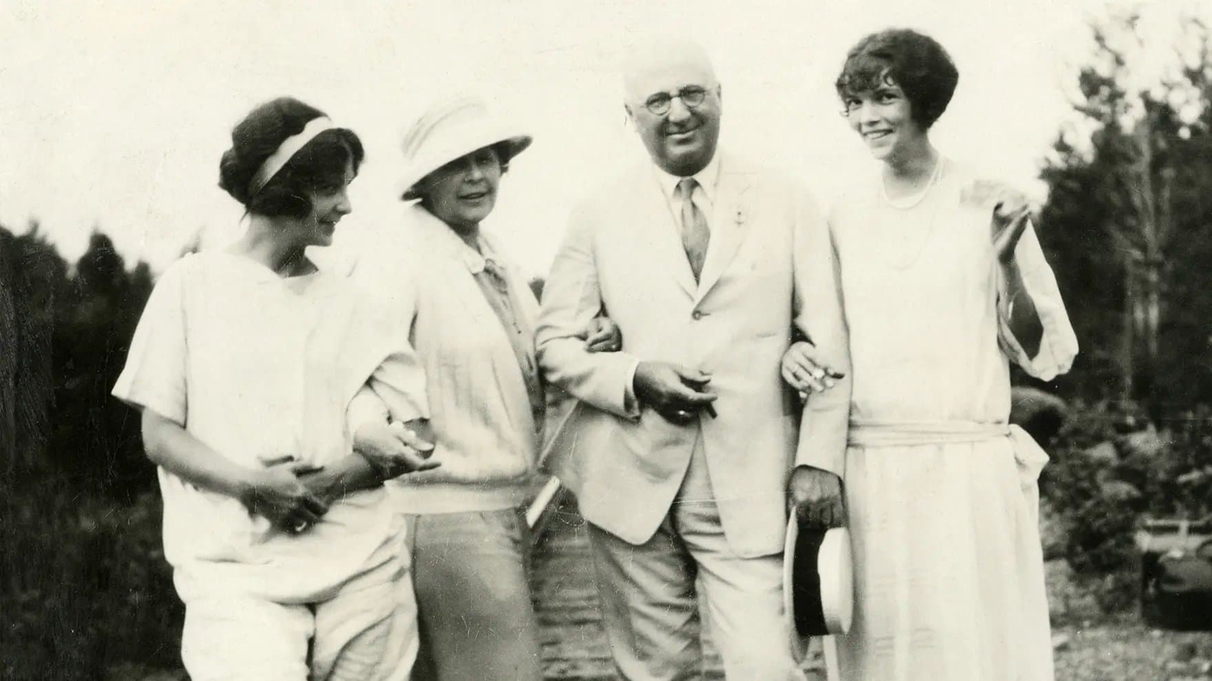 A group of three women and a man stand closely, smiling, in light-colored attire. The man holds a hat. They are outdoors with trees in the background.