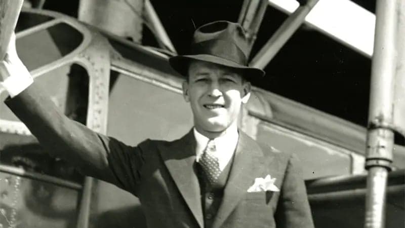 A man in a suit and fedora smiles, standing outdoors. He holds onto a metal structure, with an industrial setting and partially visible machinery in the background.