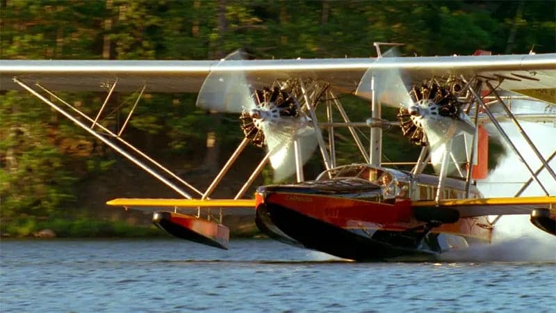A seaplane with spinning propellers skims across a calm body of water, surrounded by a forested landscape. The plane is marked with "California Clipper" on its nose.