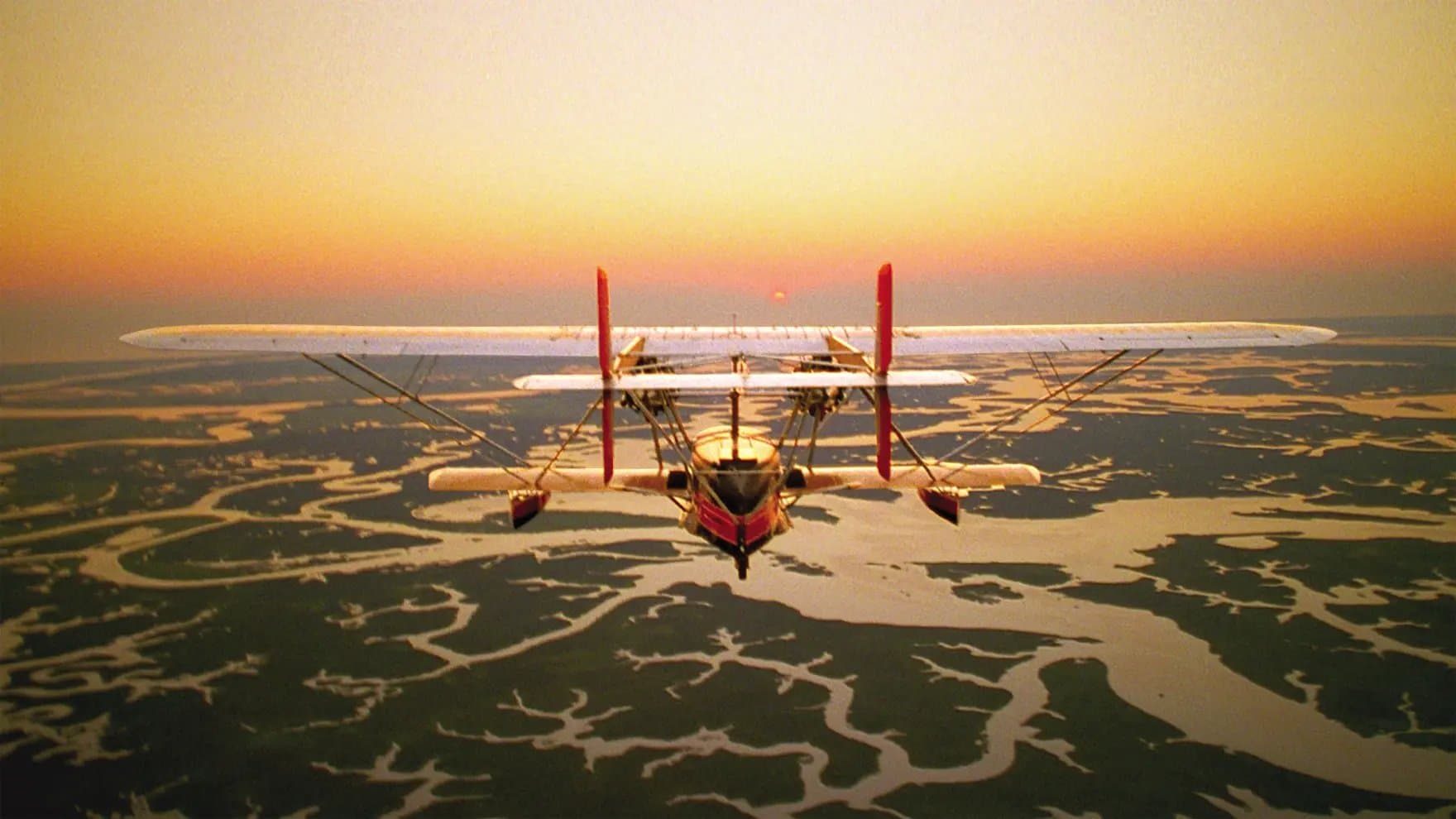 A biplane flies against a vibrant sunset backdrop, over a landscape of winding rivers and dark patches of land, creating a striking silhouette against the glowing sky.