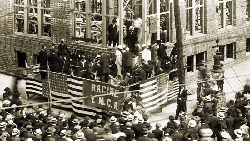 A crowd gathers near a partially constructed brick building, watching a group of people with a banner reading "RACINE Y.M.C.A." and American flags. Some individuals wear hats and coats.