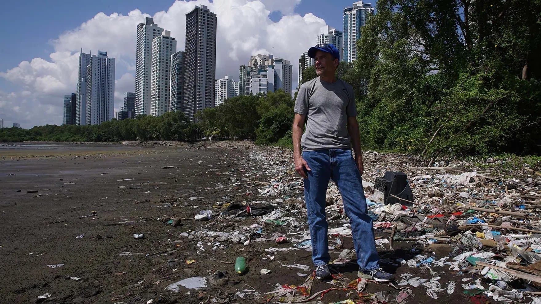 A man stands on a littered shoreline, wearing a gray shirt and blue jeans. Skyscrapers and trees form the background under a cloudy sky.