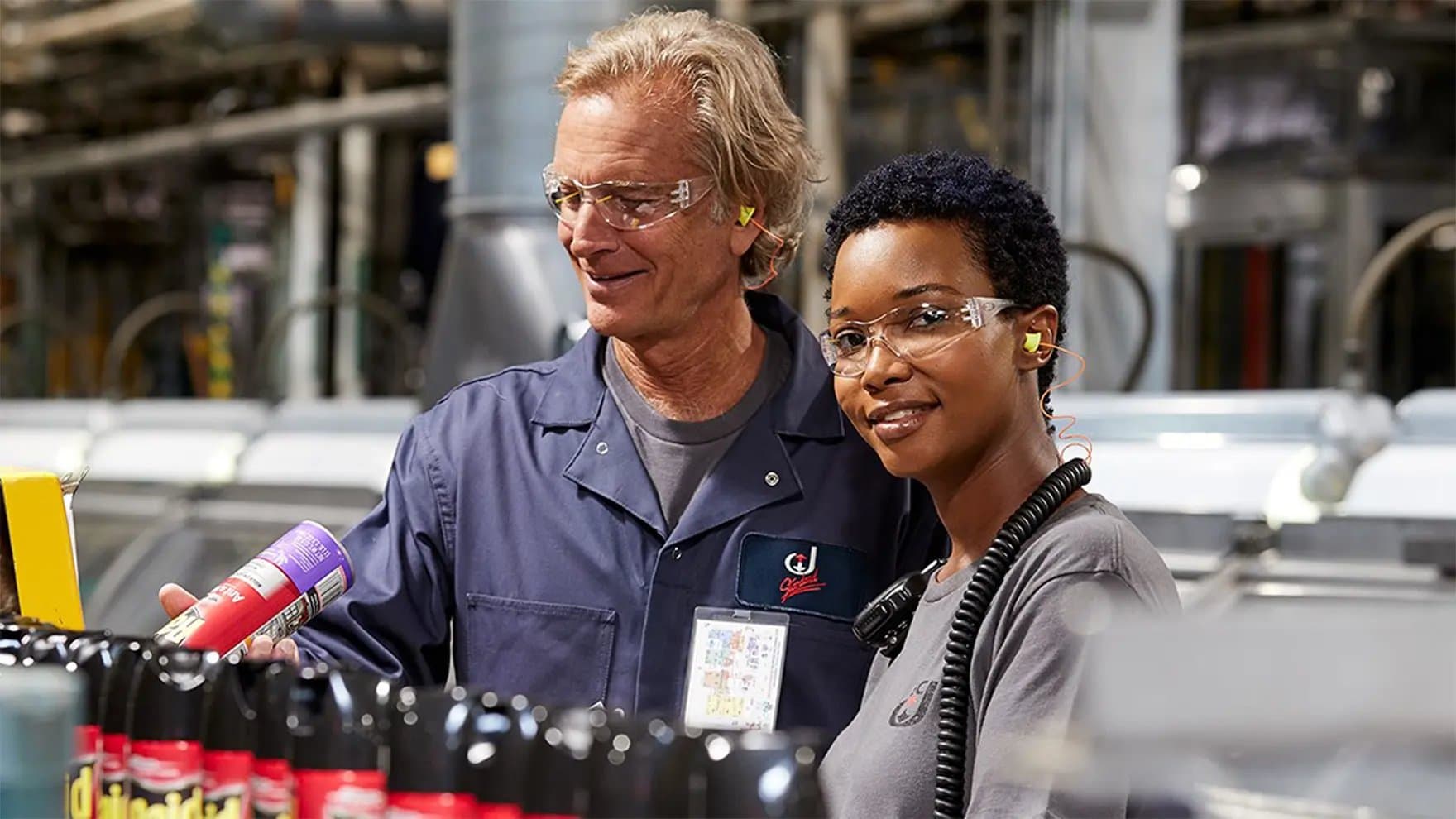Two factory workers wearing safety glasses and ear protection stand near a conveyor belt with bottles, smiling, in an industrial setting. One holds a can labeled "X-14."