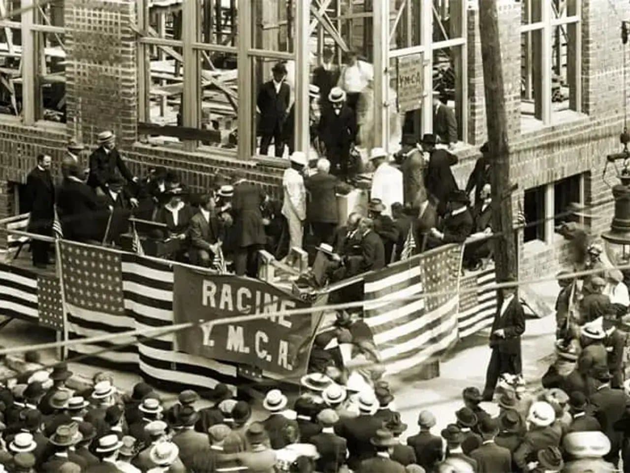 People gather around a construction site featuring American flags and a "Racine Y.M.C.A." banner. They're witnessing an event with brick buildings and scaffolding in the background.