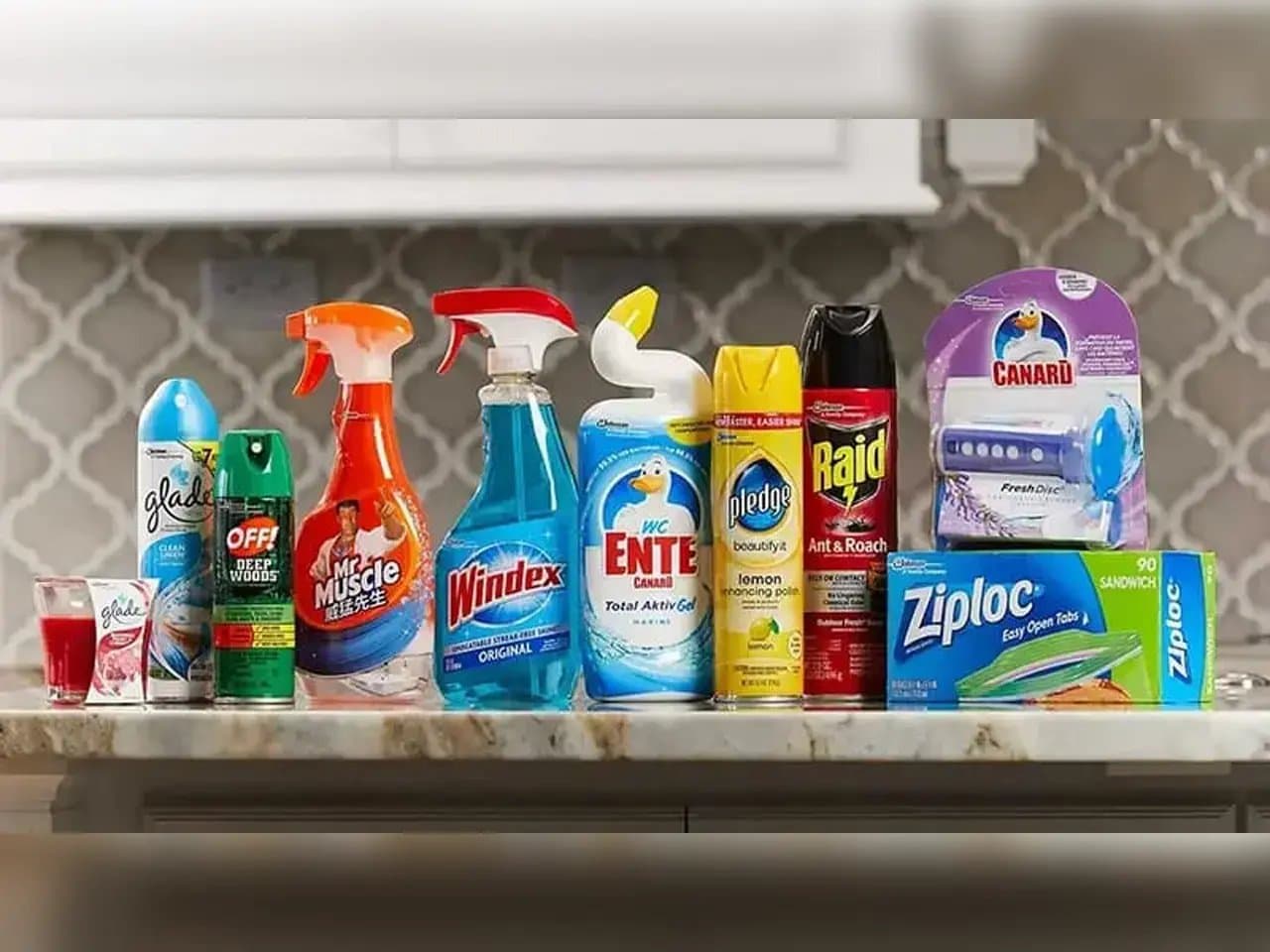 A collection of household cleaning and pest control products is lined up on a countertop. Brands visible include Glade, OFF!, Mr. Muscle, Windex, WC Ente, Pledge, Raid, Canard, and Ziploc.