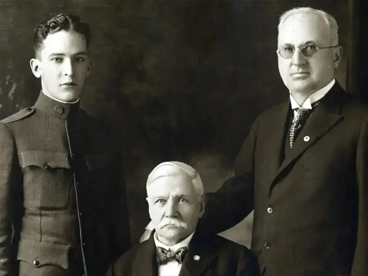 Three men are captured in a formal portrait. A young man in military uniform stands on the left, an older seated man with a mustache is in the center, and a man in a dark suit with glasses stands on the right. They are in a studio setting with a plain background.