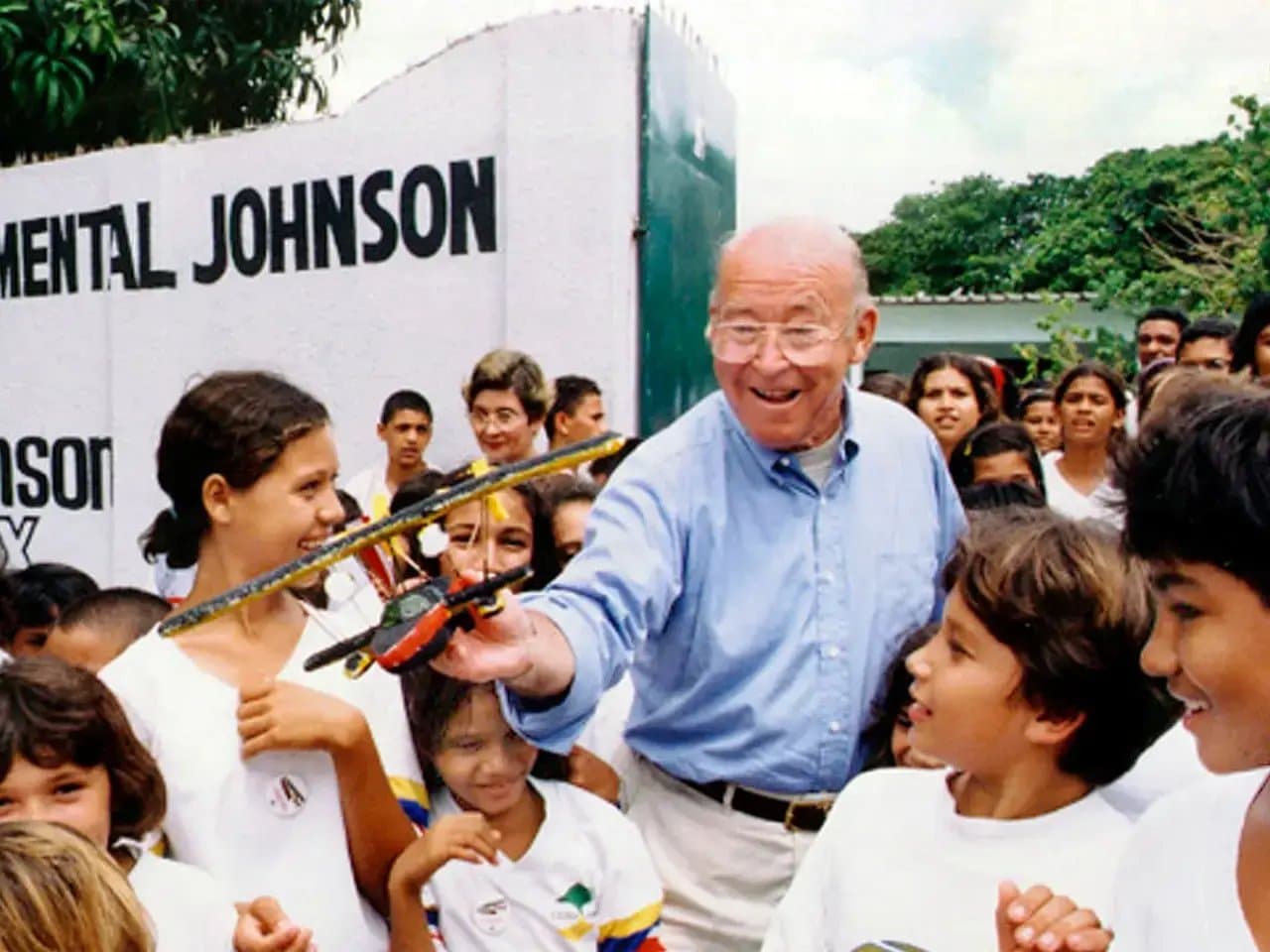 A man playfully holds a model airplane towards smiling children. They stand in front of a wall with text: “MENTAL JOHNSON.” The setting includes a leafy outdoor area with many people.