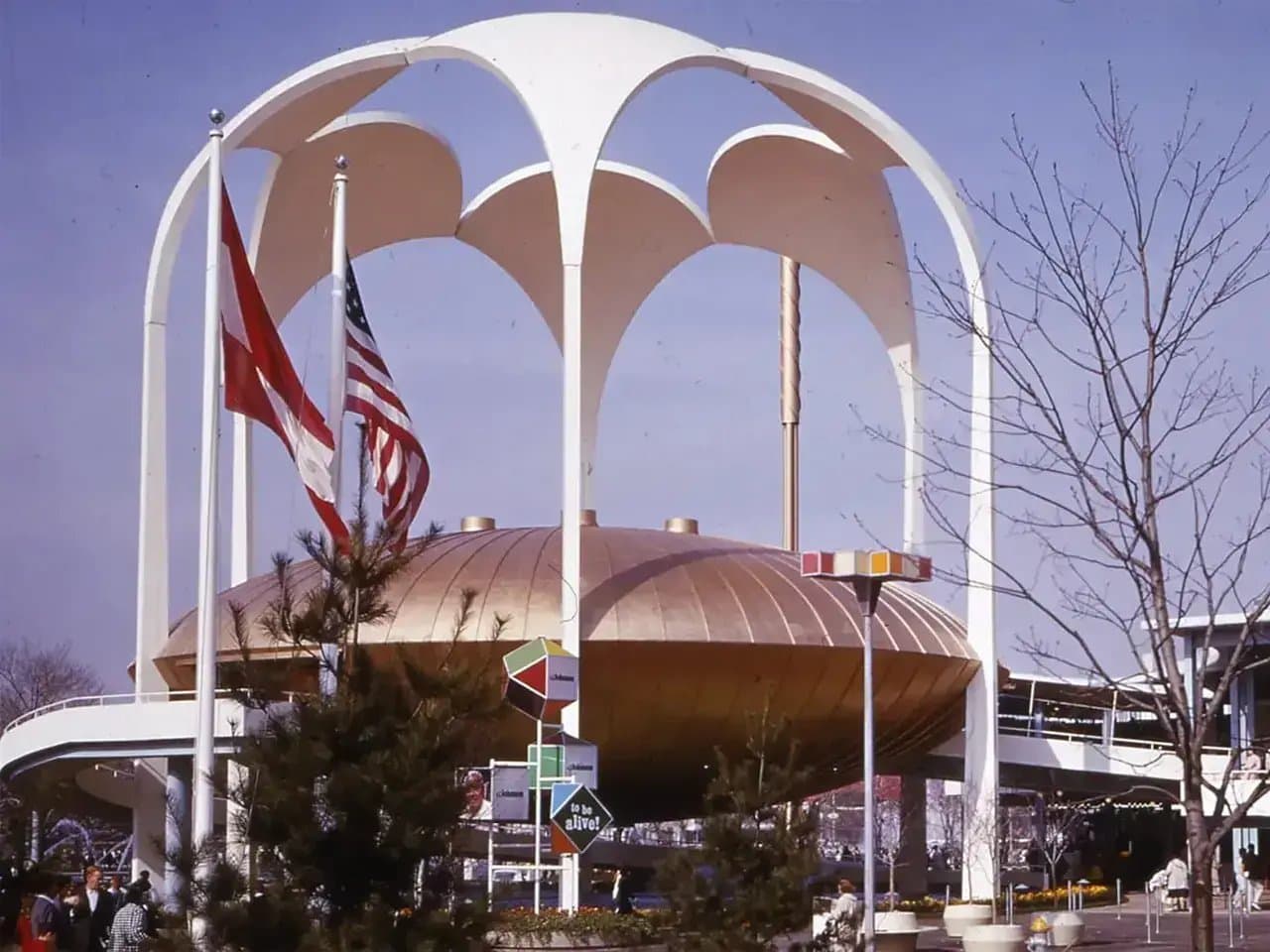 A futuristic pavilion structure with a domed roof and arching supports stands. Two flags wave beside it. Surrounding trees and signs add context to this outdoor setting.