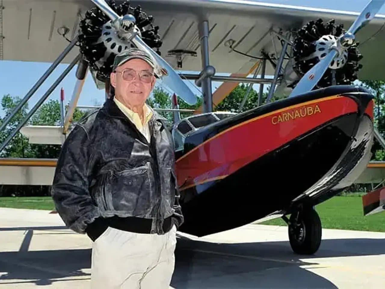 A man stands in front of a vintage biplane titled "CARNAUBA," wearing a cap and leather jacket, with trees visible in the sunny background.