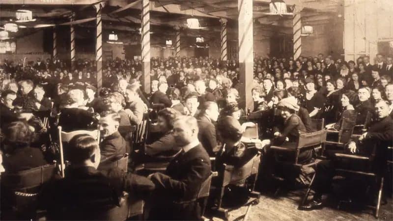 A large group of people sitting attentively in rows of chairs, facing forward, in a dimly lit, spacious hall with decorative pillars and overhead lamps.