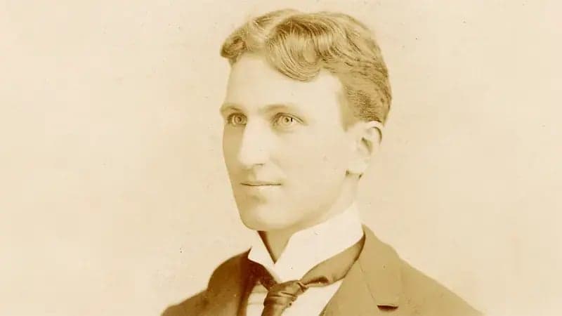 A young man stands, facing slightly left, wearing a suit with a high-collared shirt and tie. The sepia-toned portrait suggests a late 19th or early 20th-century setting.