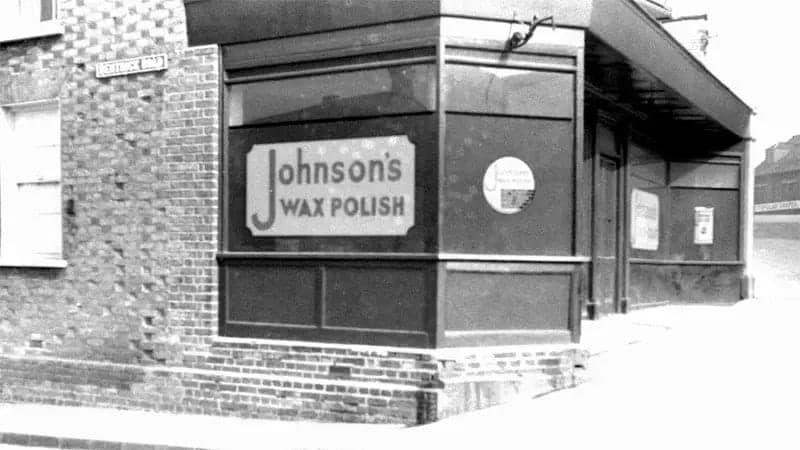 A brick building corner displays large signs reading "Johnson's Wax Polish." Surrounding it are brick walls and windows, creating a classic, early 20th-century street corner scene.