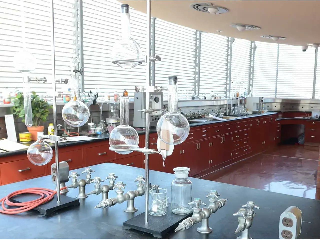 Glass laboratory apparatus sits on a countertop with multiple connected glass flasks held in clamps. Surrounding are stainless-steel faucets, red cabinets, and large windows providing ample light.