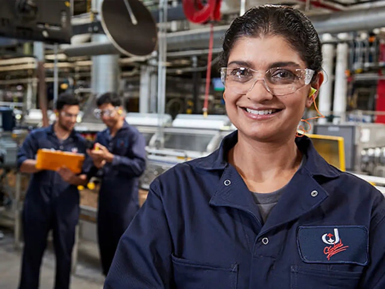 Worker smiling, wearing safety glasses and earplugs, stands confidently in a factory setting. Two colleagues behind are discussing, holding a clipboard amidst industrial machinery and pipes.